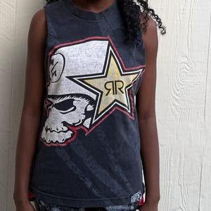 May include: Black sleeveless graphic tee featuring a skull design with a star and the letters "RR". The design is in white, gold, and red on a dark gray background. The shirt has a distressed look.