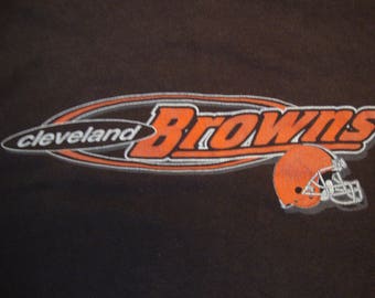 Puede incluir: Una camiseta negra con un logotipo de los Cleveland Browns en blanco y naranja. El logotipo incluye el nombre del equipo "Browns" y la ciudad "Cleveland" en un contorno blanco. Un casco de fútbol marrón está incluido en el logotipo.