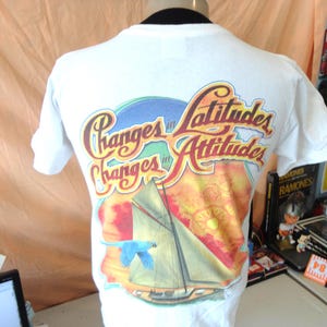 Pode incluir: T-shirt branco com um gráfico colorido nas costas. O gráfico apresenta um veleiro, um pássaro azul e as palavras "Changes in Latitudes, Changes in Attitudes" em uma fonte estilizada. A camisa tem mangas curtas.