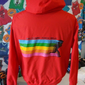 May include: A red hooded jacket with long sleeves. The back of the jacket features a colorful graphic with the text "1984 Olympic Torch Relay". The graphic includes a rainbow of colors and a black outline.