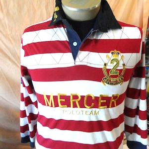 May include: A long-sleeve rugby shirt featuring horizontal red and white stripes, a black collar, and a blue placket with a single white button. The chest displays the gold embroidered text MERCER POLOTEAM and a large gold crest with the number 2.