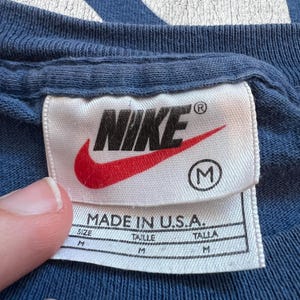 Puede incluir: Una etiqueta blanca con el logotipo de Nike en rojo y negro, el texto "MADE IN U.S.A." y la talla "M" en un c&iacute;rculo. La etiqueta est&aacute; unida a una camisa azul.