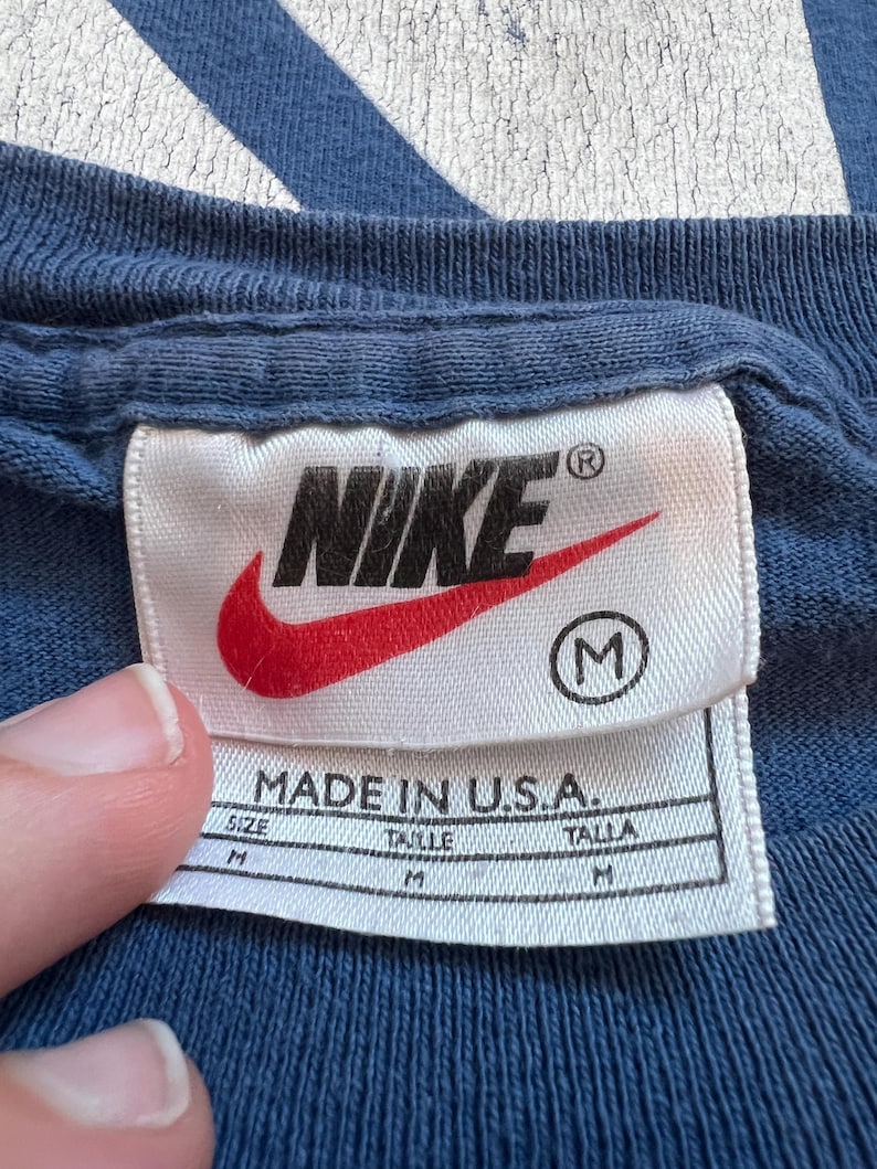 Puede incluir: Una etiqueta blanca con el logotipo de Nike en rojo y negro, el texto "MADE IN U.S.A." y la talla "M" en un c&iacute;rculo. La etiqueta est&aacute; unida a una camisa azul.