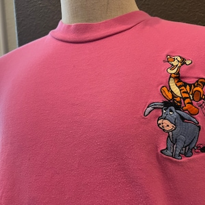 Può includere: Maglietta rosa a maniche lunghe con un'immagine ricamata di Tigro e Ih-Oh di Winnie the Pooh.