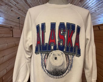 May include: A white sweatshirt with a large graphic print of the word "ALASKA" in red, blue, and gray letters. The graphic also includes a polar bear with the words "Truth, Wisdom, Knowledge" around it.