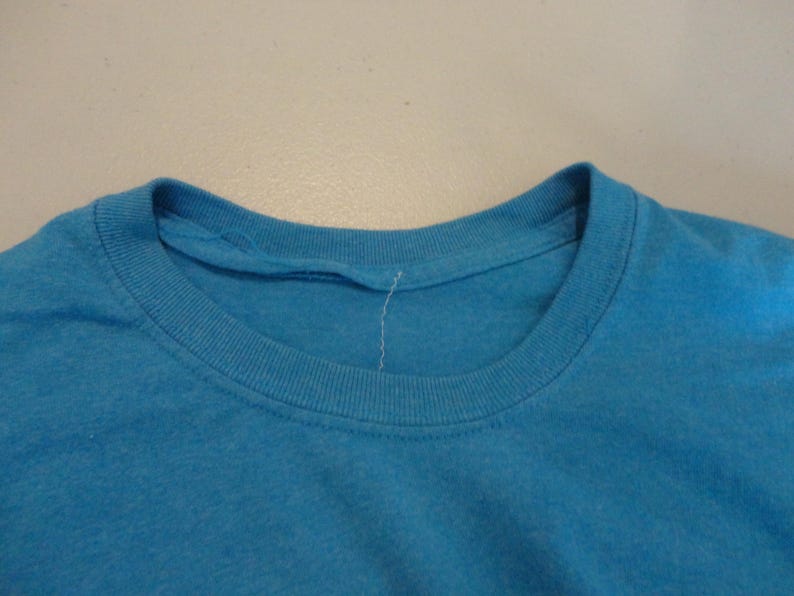 May include: A close-up shot of a turquoise crew neck t-shirt. The neckline is ribbed and the fabric appears soft. The shirt is a solid colour and has a simple, classic design.