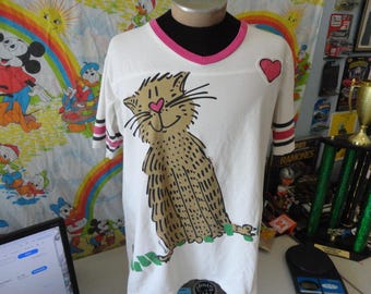 May include: White t-shirt with a cartoon cat illustration. The cat has a pink heart nose and is sitting on green grass. The shirt has pink trim on the collar and sleeves, with a red heart patch.
