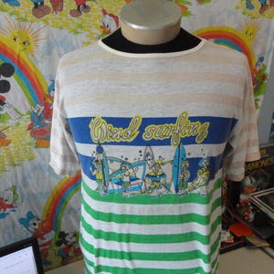 May include: A striped t-shirt with the words "Wind Surfing" in yellow script across the chest. The shirt features a blue band with cartoon surfers and surfboards, and green and white stripes at the bottom.