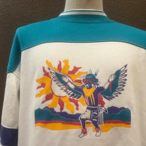 Puede incluir: Sudadera blanca con un diseño de bloques de color turquesa y morado. La parte delantera presenta un gráfico de una figura nativa americana estilizada con alas, bailando frente a un sol.