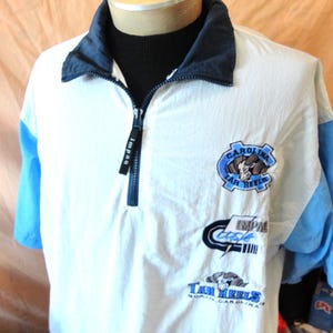 Puede incluir: Un su&eacute;ter blanco de manga corta con cuello de cremallera de un cuarto en azul oscuro y mangas azul claro. El frente presenta logotipos bordados que incluyen una mascota de carnero, el texto Carolina Tar Heels y la marca Impaq en el tirador.