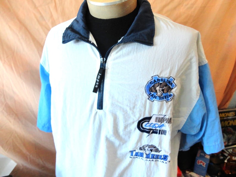 Puede incluir: Un su&eacute;ter blanco de manga corta con cuello de cremallera de un cuarto en azul oscuro y mangas azul claro. El frente presenta logotipos bordados que incluyen una mascota de carnero, el texto Carolina Tar Heels y la marca Impaq en el tirador.