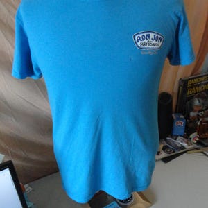 May include: A turquoise t-shirt with a round neck and short sleeves. The shirt displays a logo reading "RON JON SURFBOARDS" in a white and blue design on the upper left chest.