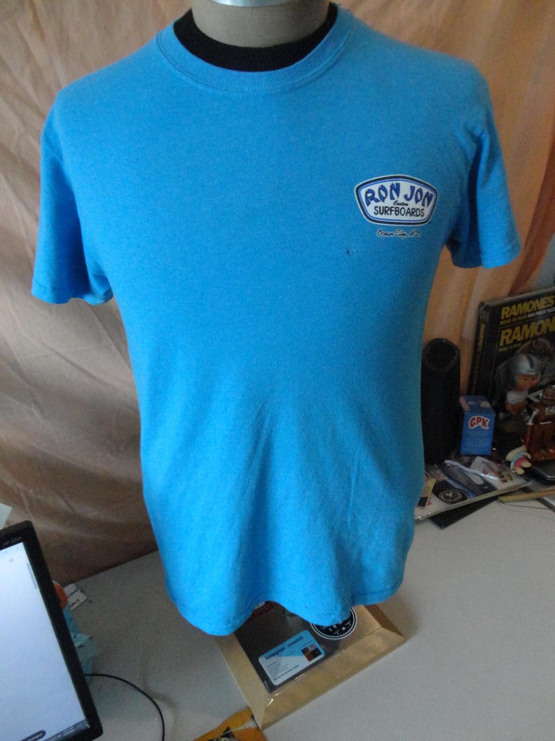 May include: A turquoise t-shirt with a round neck and short sleeves. The shirt displays a logo reading "RON JON SURFBOARDS" in a white and blue design on the upper left chest.