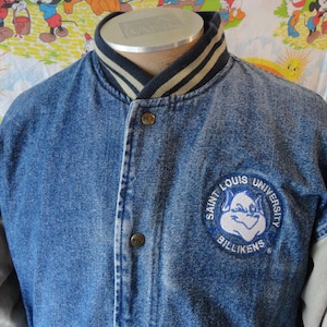 May include: A blue denim jacket with gray sleeves and a striped collar. The jacket features a Saint Louis University Billikens patch on the chest. The jacket has button closures and a classic varsity style.