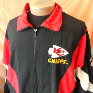 Pode incluir: Casaco preto com z&iacute;per e detalhes em vermelho e branco. Apresenta o logotipo dos Kansas City Chiefs no peito, com a palavra "CHIEFS" em amarelo. O casaco tem gola vermelha e &eacute; exibido em um manequim.