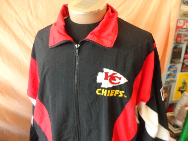 Pode incluir: Casaco preto com z&iacute;per e detalhes em vermelho e branco. Apresenta o logotipo dos Kansas City Chiefs no peito, com a palavra "CHIEFS" em amarelo. O casaco tem gola vermelha e &eacute; exibido em um manequim.