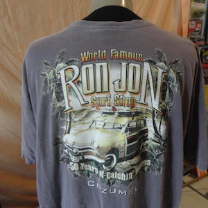 May include: A gray t-shirt featuring the text "World Famous Ron Jon Surf Shop" with a vintage car graphic. The design includes palm trees and the text "50 Years of Catchin' Waves Cozumel".