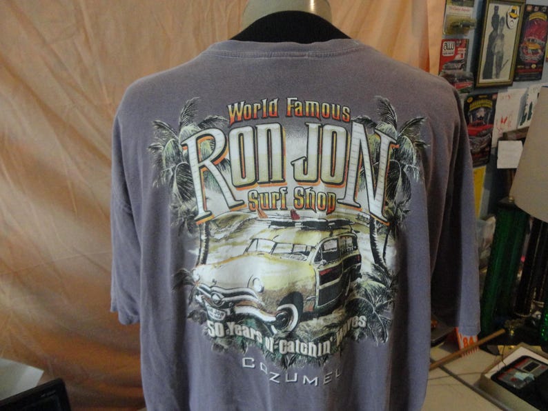 May include: A gray t-shirt featuring the text "World Famous Ron Jon Surf Shop" with a vintage car graphic. The design includes palm trees and the text "50 Years of Catchin' Waves Cozumel".
