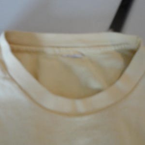 May include: Close-up of a cream-coloured t-shirt neckline. The collar is a darker shade of cream than the body of the shirt. The fabric appears to be soft and slightly worn.