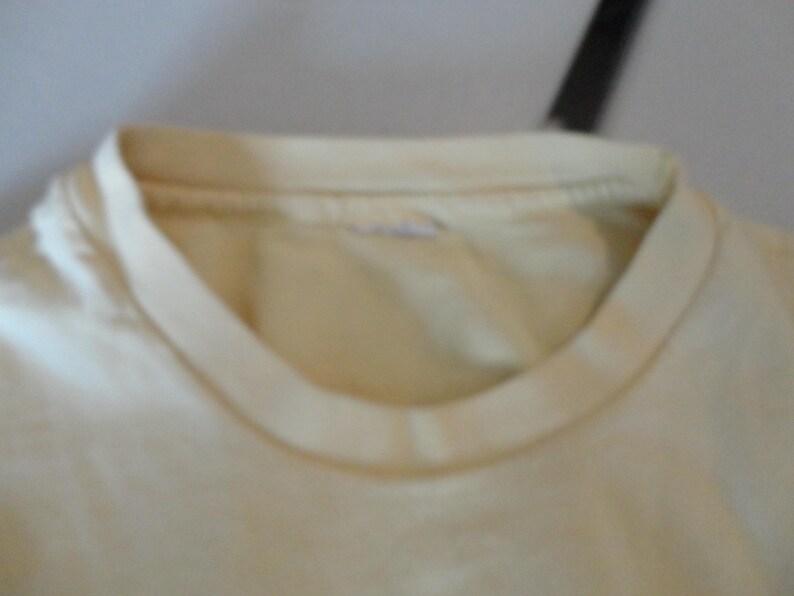 May include: Close-up of a cream-coloured t-shirt neckline. The collar is a darker shade of cream than the body of the shirt. The fabric appears to be soft and slightly worn.