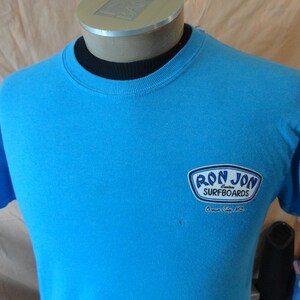 May include: A turquoise t-shirt featuring the "Ron Jon Surfboards" logo. The logo is in a white and blue design. The shirt has short sleeves and a crew neck.