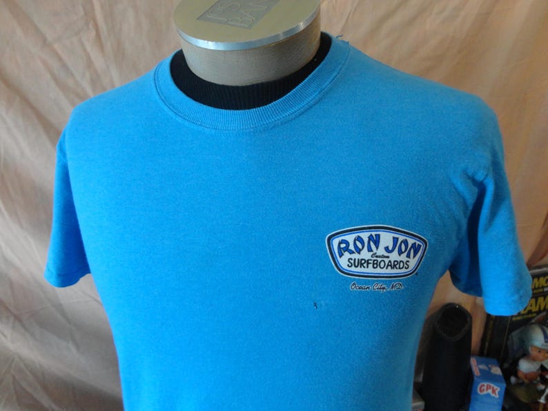 May include: A turquoise t-shirt featuring the "Ron Jon Surfboards" logo. The logo is in a white and blue design. The shirt has short sleeves and a crew neck.