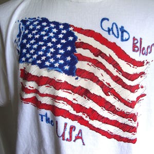 Puede incluir: Camiseta blanca con un diseño desgastado de la bandera estadounidense en rojo, blanco y azul. Las palabras "GOD Bless" y "The USA" están impresas encima y debajo del gráfico de la bandera.