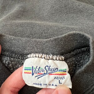May include: A gray sweatshirt tag with the text "Velva Sheen" in a rainbow font. The tag also says "Adult L" and "Made in U.S.A."