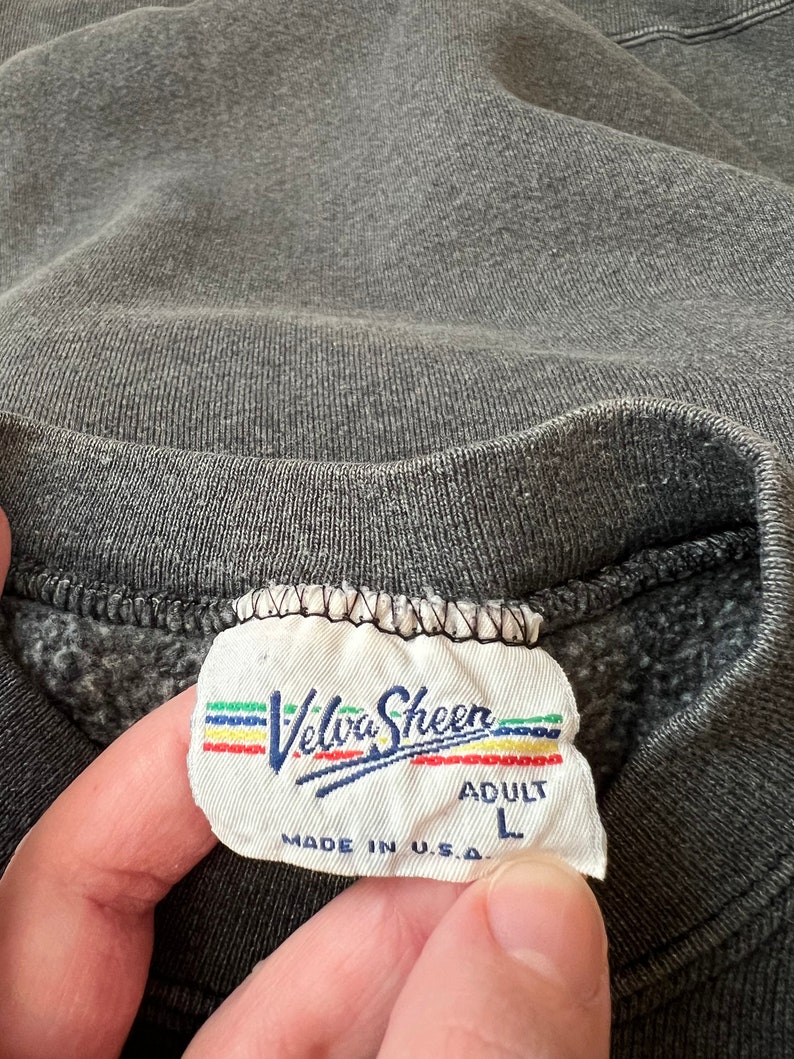 May include: A gray sweatshirt tag with the text "Velva Sheen" in a rainbow font. The tag also says "Adult L" and "Made in U.S.A."