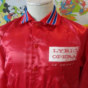 May include: A red satin jacket with a white rectangular patch that reads "LYRIC OPERA OF CHICAGO." The collar is trimmed with blue and white stripes. The jacket has red buttons and a classic design.