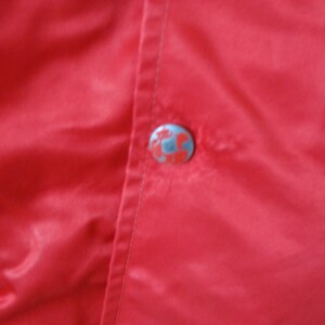 May include: A close-up of a red satin jacket with a single button closure. The button is silver with a design on it.