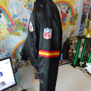 May include: Black satin bomber jacket with the NFL logo on the sleeve. The jacket has red and yellow stripes on the sleeve and a white patch on the chest. The jacket is hanging on a white surface.