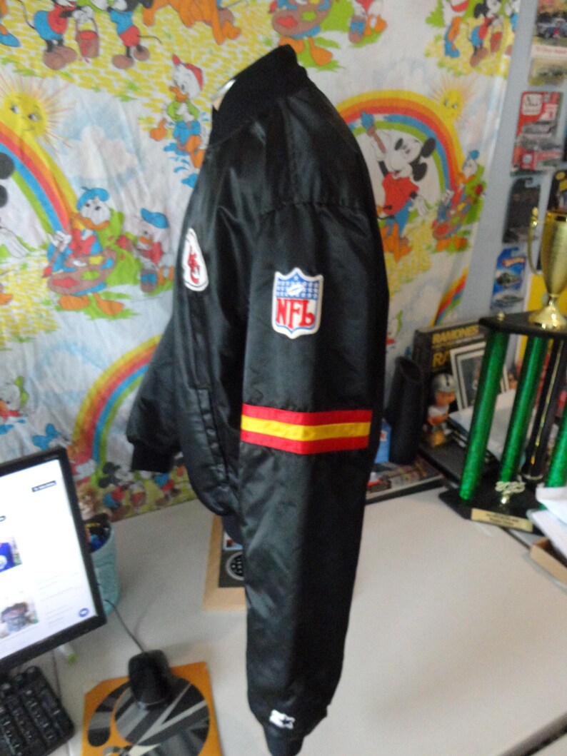 May include: Black satin bomber jacket with the NFL logo on the sleeve. The jacket has red and yellow stripes on the sleeve and a white patch on the chest. The jacket is hanging on a white surface.