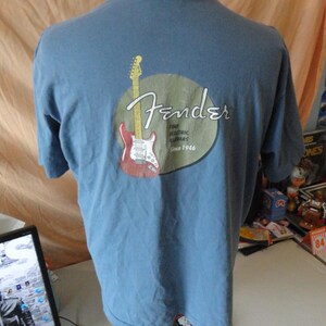 May include: A light blue t-shirt featuring a Fender logo with a red and white guitar graphic. The logo is inside a tan circle, with the words "Fender Fine Electric Guitars Since 1946".