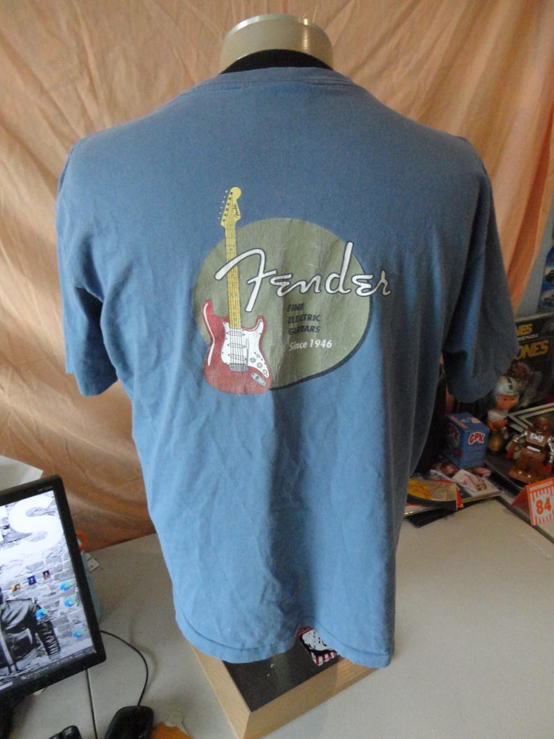 May include: A light blue t-shirt featuring a Fender logo with a red and white guitar graphic. The logo is inside a tan circle, with the words "Fender Fine Electric Guitars Since 1946".