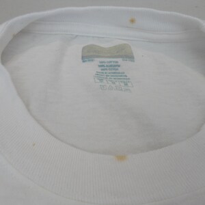 May include: Close-up of a white crew neck t-shirt. The shirt has a tag with text and care instructions. There are a few small stains on the shirt. The collar is ribbed.