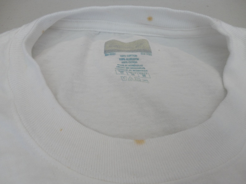 May include: Close-up of a white crew neck t-shirt. The shirt has a tag with text and care instructions. There are a few small stains on the shirt. The collar is ribbed.