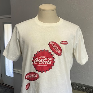 May include: White t-shirt with a red Coca-Cola logo and several red Coca-Cola bottle caps printed on it. The text on the logo reads "Enjoy Coca-Cola Trademark®".