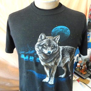 May include: A black short-sleeve t-shirt featuring a graphic print of a wolf standing in a landscape at night. The design includes a large blue moon in the sky, a dark forest silhouette, and blue light accents on the ground beneath the wolf.