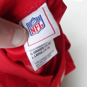 以下が含まれることがあります： NFLのロゴが入った白いタグが付いた赤い衣服のクローズアップ。タグには「G GRANDE (14-16)」と「L LARGE (14-16)」のサイズが記載されています。タグの洗濯表示には「TURN GARMENT INSIDE OUT」と「MACHINE WASH COLD SEPARATELY」と記載されています。