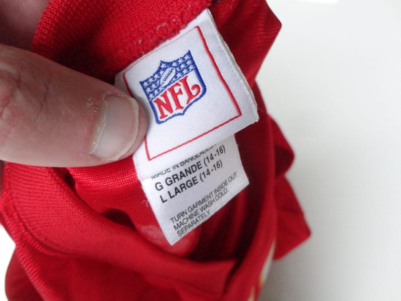 以下が含まれることがあります： NFLのロゴが入った白いタグが付いた赤い衣服のクローズアップ。タグには「G GRANDE (14-16)」と「L LARGE (14-16)」のサイズが記載されています。タグの洗濯表示には「TURN GARMENT INSIDE OUT」と「MACHINE WASH COLD SEPARATELY」と記載されています。