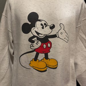 May include: A light gray sweatshirt featuring a large graphic of Mickey Mouse. Mickey is depicted in his classic pose, with black ears, a black body, red shorts, and yellow shoes. The sweatshirt has a speckled pattern.