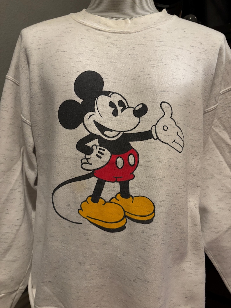 May include: A light gray sweatshirt featuring a large graphic of Mickey Mouse. Mickey is depicted in his classic pose, with black ears, a black body, red shorts, and yellow shoes. The sweatshirt has a speckled pattern.