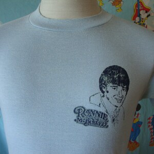May include: Light blue t-shirt with a vintage print of Ronnie McDowell. The print features a black and white image of Ronnie McDowell with the text "Ronnie McDowell" underneath.