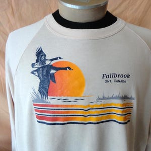 May include: Beige sweatshirt with a graphic of two geese flying in front of a sunset. The design includes orange, yellow, and red stripes, and the text "Fallbrook ONT CANADA".