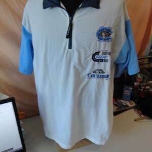 Puede incluir: Un polo blanco de manga corta con mangas azul claro y cuello azul oscuro. La prenda cuenta con una cremallera frontal de un cuarto con un tirador negro. Tres logotipos distintos est&aacute;n impresos en el pecho, incluyendo el texto Tar Heels.