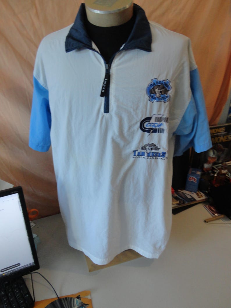 Puede incluir: Un polo blanco de manga corta con mangas azul claro y cuello azul oscuro. La prenda cuenta con una cremallera frontal de un cuarto con un tirador negro. Tres logotipos distintos est&aacute;n impresos en el pecho, incluyendo el texto Tar Heels.