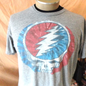 May include: Gray t-shirt featuring a colorful graphic of a skull with a lightning bolt and tie-dye design in red, white, and blue. The shirt has a crew neck and short sleeves. The design is a popular symbol.