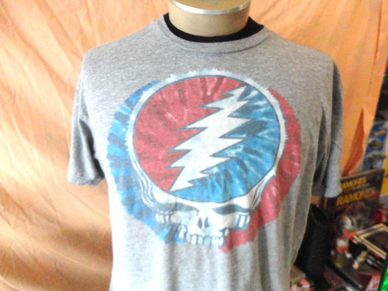 May include: Gray t-shirt featuring a colorful graphic of a skull with a lightning bolt and tie-dye design in red, white, and blue. The shirt has a crew neck and short sleeves. The design is a popular symbol.