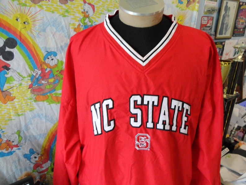 May include: Red windbreaker jacket with white NC STATE lettering and a red and white logo. The jacket has a black and white striped collar.